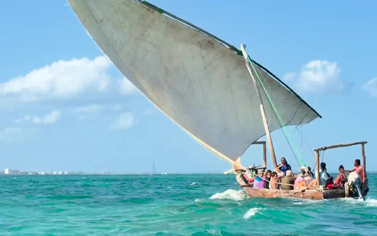 A dhow under a white lateen sail, crossing turquoise water